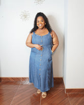 Plus size model wearing a medium wash denim midi dress with adjustable straps, button front detail, smocked back, and side pockets.