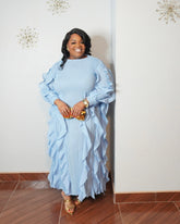 Plus size model wearing a sky blue pleated maxi dress with cascading ruffle details and gold accessories.