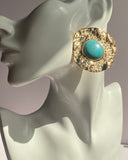 Gold textured statement earrings featuring a turquoise center stone displayed on an earring stand.