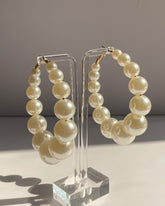 Graduated ivory pearl hoop earrings with gold-tone hardware displayed on a clear acrylic stand.
