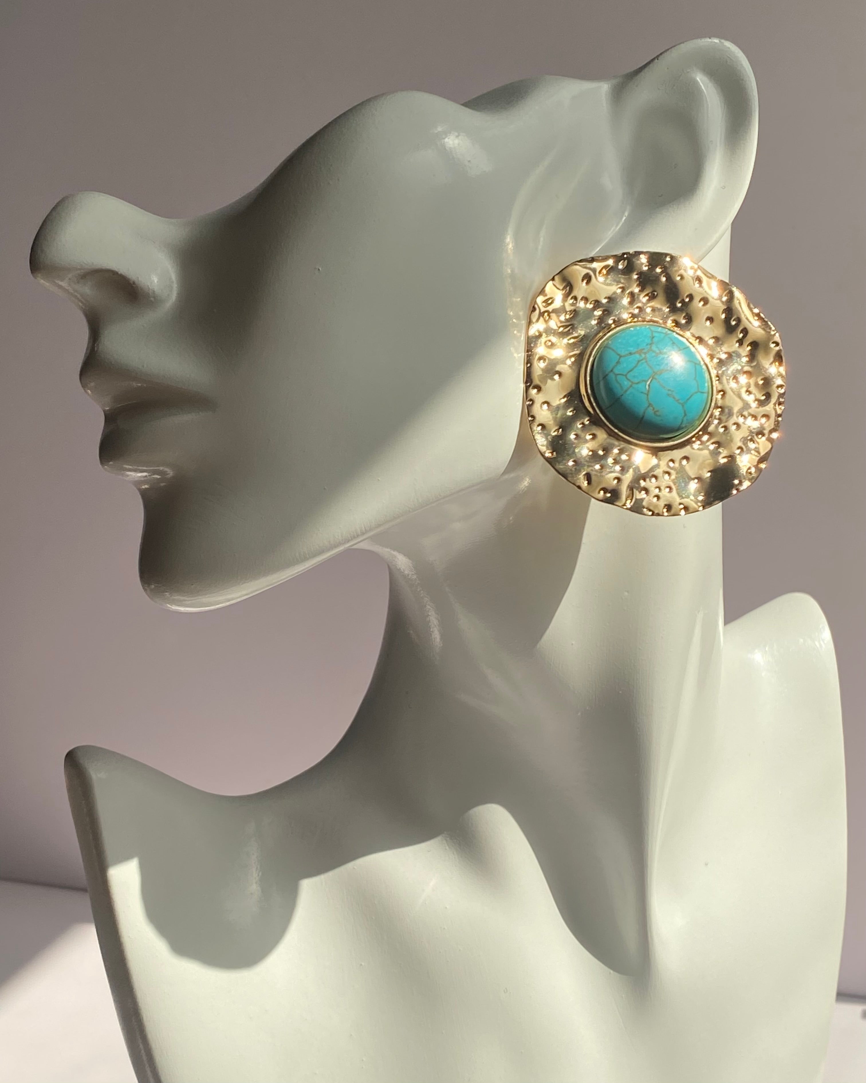 Gold textured statement earrings featuring a turquoise center stone displayed on an earring stand.