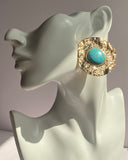 Gold textured statement earrings featuring a turquoise center stone displayed on an earring stand.
