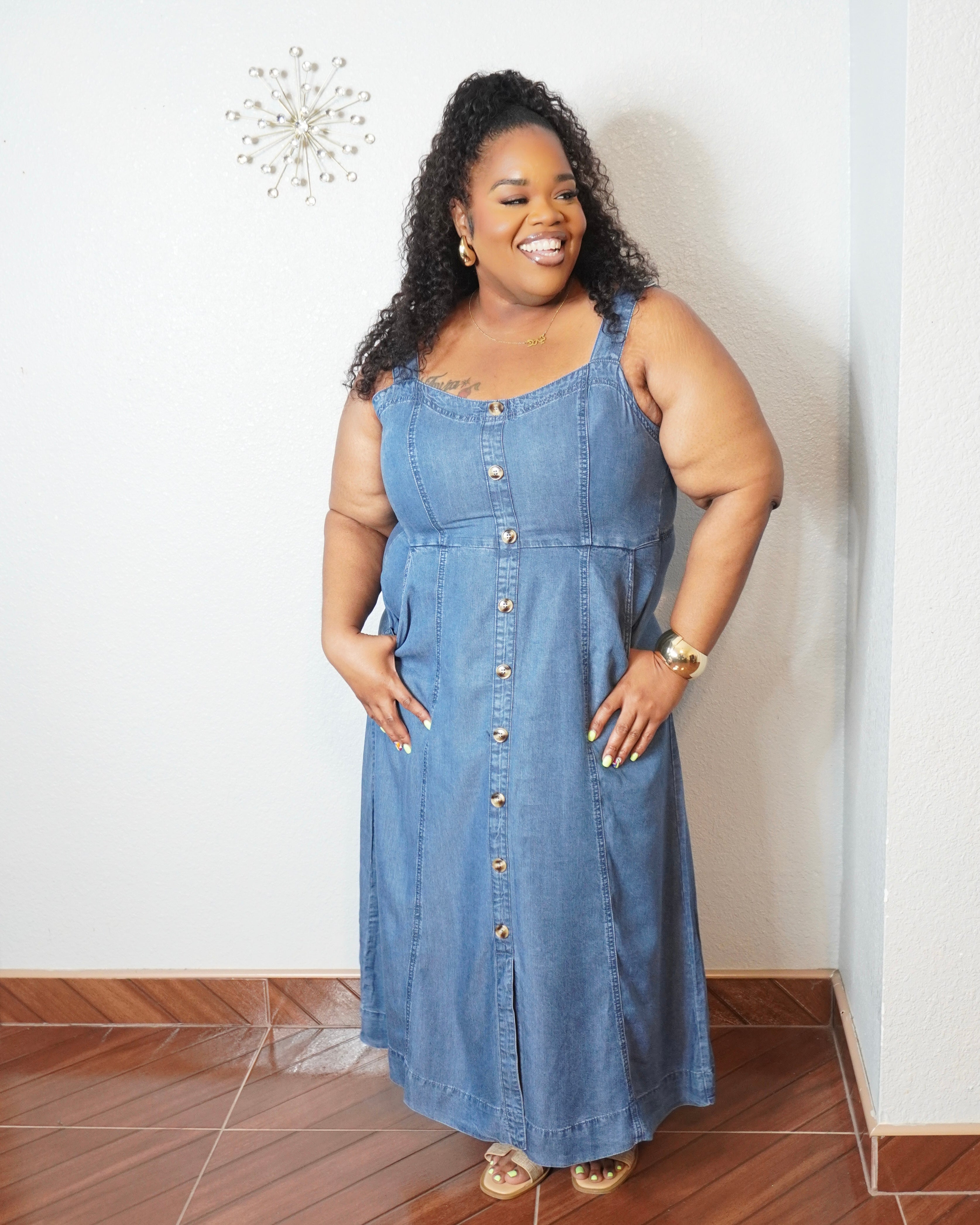 Plus size model wearing a medium wash denim midi dress with adjustable straps, button front detail, smocked back, and side pockets.