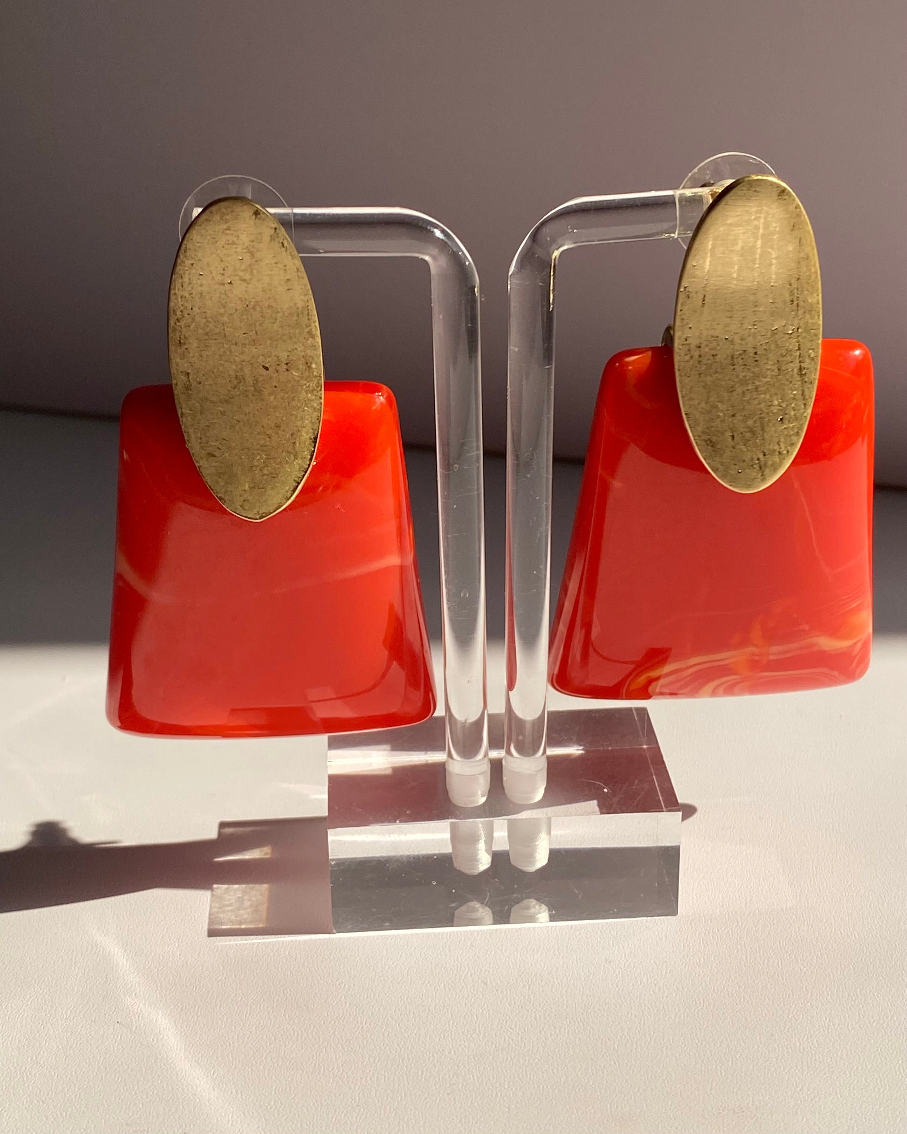 Modern resin statement earrings featuring brushed gold oval studs with glossy coral red geometric drops.