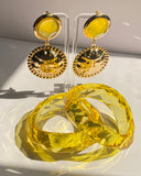 A pair of gold-tone dangle earrings with oval yellow stones and round beaded gold pendants, displayed on clear acrylic stands above a large faceted yellow crystal-like object on a white surface.