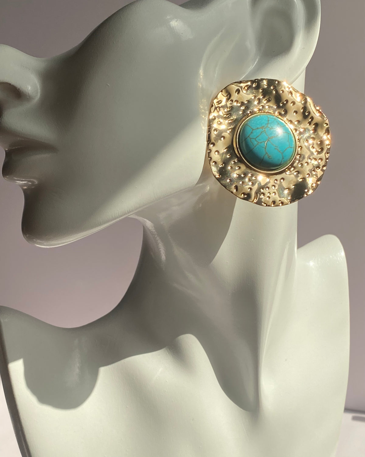 Gold textured statement earrings featuring a turquoise center stone displayed on an earring stand.