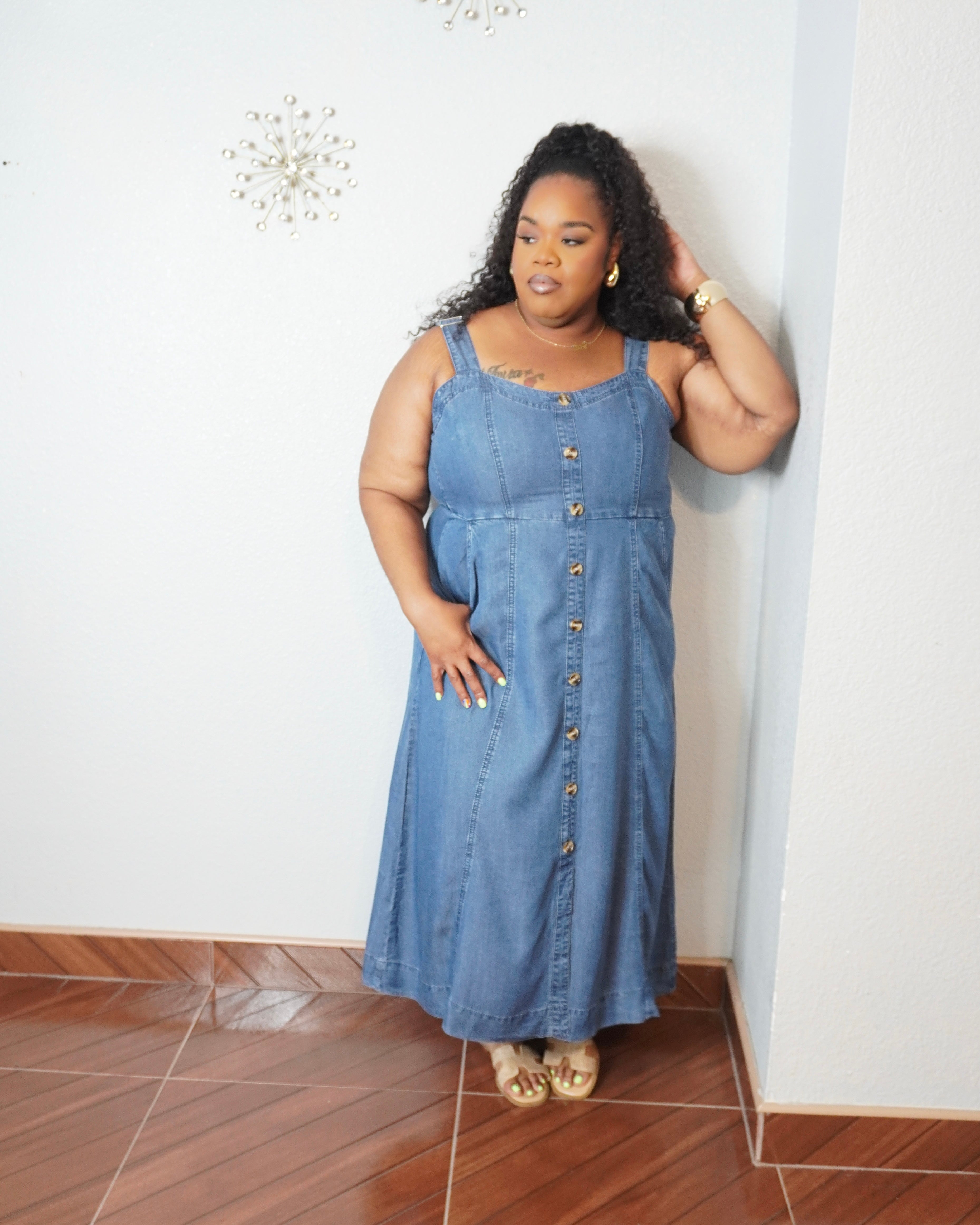 Plus size model wearing a medium wash denim midi dress with adjustable straps, button front detail, smocked back, and side pockets.
