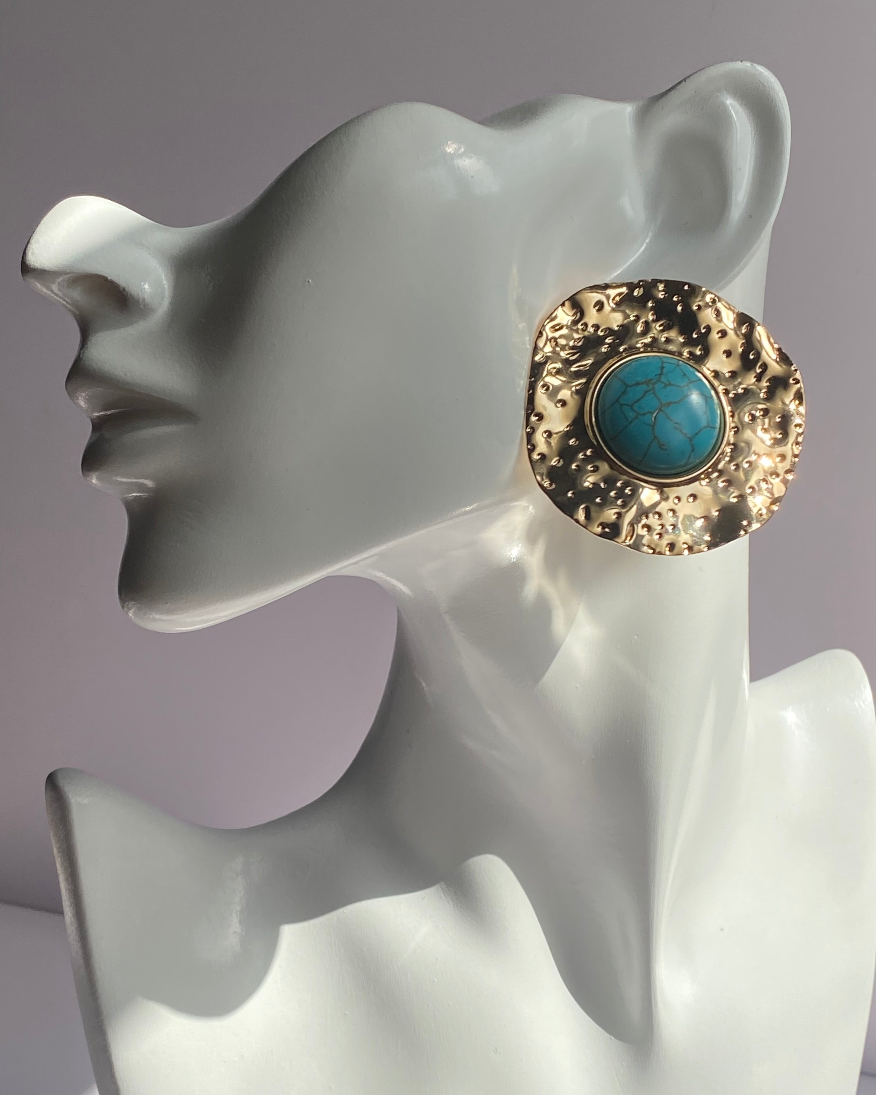 Gold textured statement earrings featuring a turquoise center stone displayed on an earring stand.