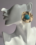 Gold textured statement earrings featuring a turquoise center stone displayed on an earring stand.