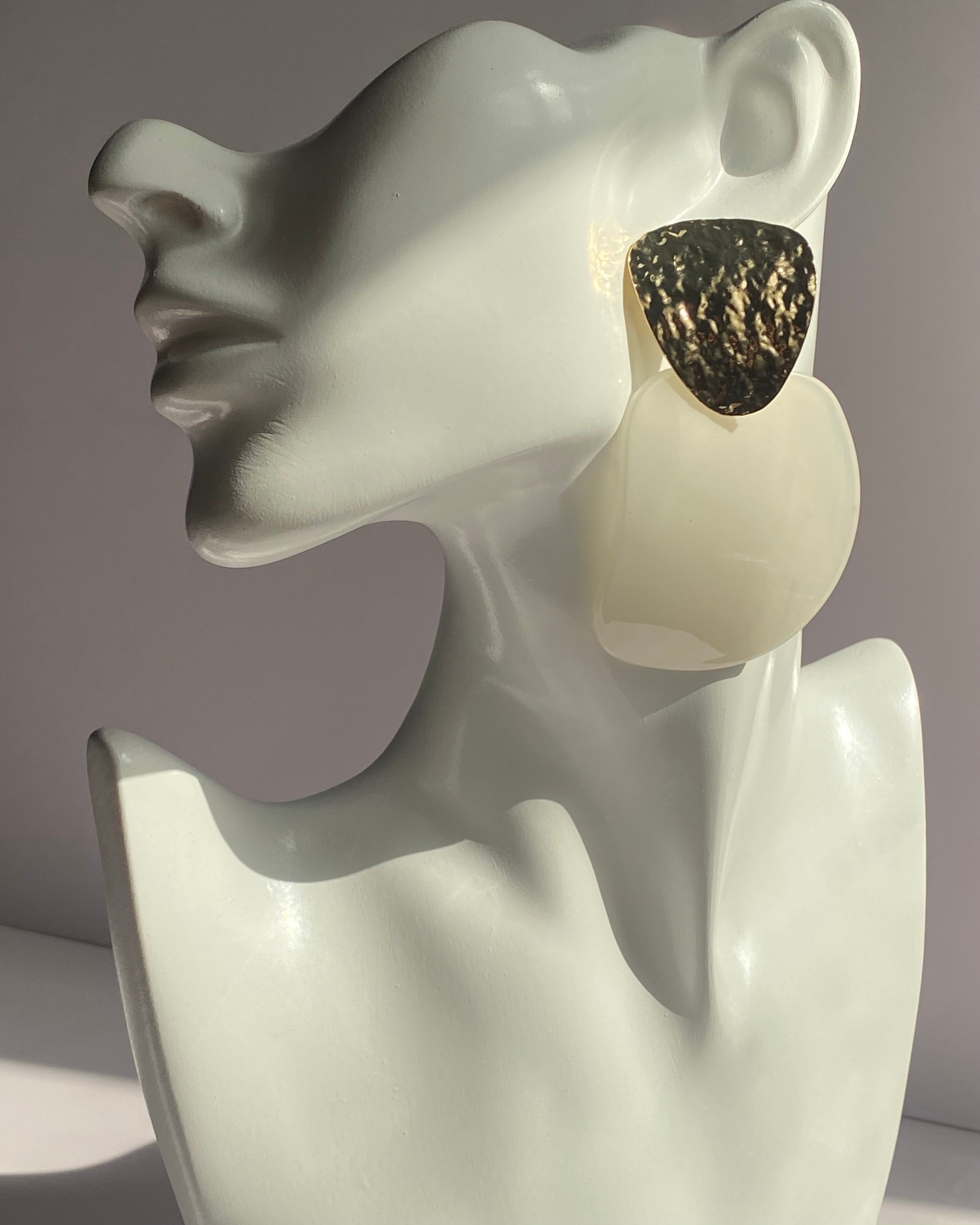 Large statement earrings displayed on a white ceramic female bust. The earrings feature a textured, hammered dark metallic top half and a smooth, opaque cream-colored bottom half.